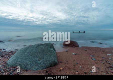 Paysage marin calme avec de gros rochers côtiers sur une plage de sable sous un ciel doux et nuageux, avec un petit épave lointaine sur l'eau immobile. Nature côtière paisible Banque D'Images