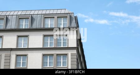Façade moderne grise d'un bâtiment à plusieurs étages avec des fenêtres blanches et un toit mansardé en métal sous un ciel bleu, vaste architecture urbaine extérieure panoramique avec Banque D'Images