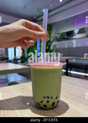 Thé à bulles au matcha avec perles de tapioca sur la table du café tandis qu'une main place une paille large dans la boisson. Culture café, tendance alimentaire, style de vie, rafraîchissement Banque D'Images