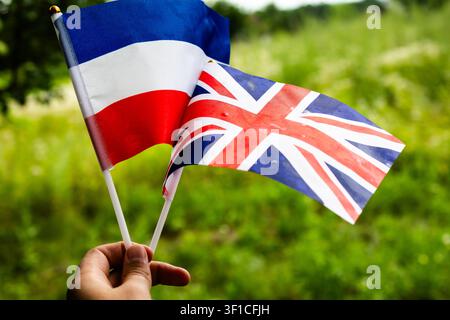 Petites drapeaux de France et du Royaume-Uni tenus ensemble dans une main contre un paysage vert doux. Le tissu lumineux qui ondule dans une brise légère suggère Banque D'Images