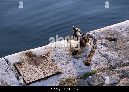 Bordure de quai en béton patiné comportant un anneau d'amarrage en métal rouillé et une plaque boulonnée, s'étendant au-dessus de l'eau calme et bleu profond de l'océan Banque D'Images