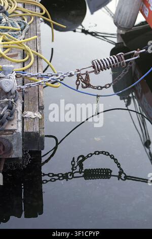 Quai maritime montrant un agencement complexe de cordes jaunes, de câbles bleus et de chaînes métalliques rouillées avec un mécanisme à ressort, se reflétant sur l'eau calme et sombre Banque D'Images