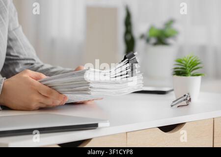 Personne tenant une grande pile de documents avec des pinces à classeur sur un bureau blanc, papier administratif et gestion, la scène comprend un ordinateur portable et un pot Banque D'Images