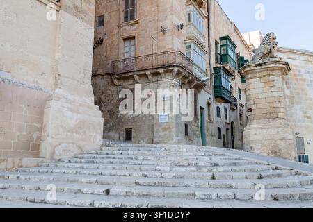De larges marches en pierre mènent à un bâtiment d'angle avec des balcons aux volets verts et la statue du lion de De Vilhena sur East Street, à La Valette, Malte Banque D'Images