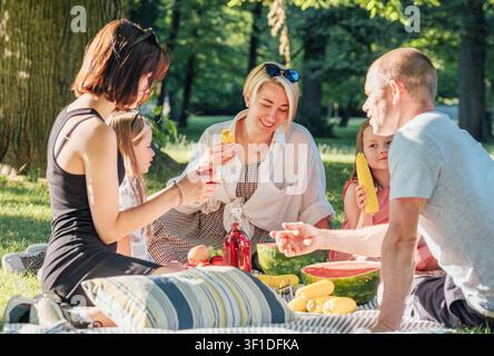 Famille et amis heureux profitant d'un pique-nique relaxant dans un parc luxuriant en été ensoleillé, assis sur une couverture douillette sur l'herbe en mangeant de la pastèque fraîche et sucrée et du jaune co Banque D'Images
