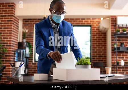 Gestionnaire organisant ses affaires sur le bureau, mettant en avant un gestionnaire masculin retirant un cahier d'un carton blanc et rangeant son bureau, homme noir en tenue de travail debout dans une pièce aux murs de briques. Banque D'Images