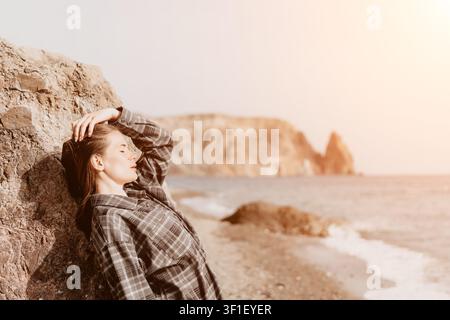 Femme détente côtière Jeune femme en chemise à carreaux s'appuie contre un rocher sur une plage ensoleillée en profitant de la brise marine Banque D'Images