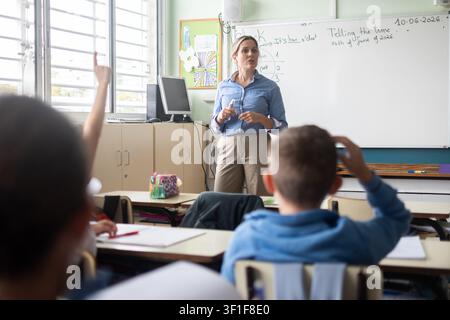 Vue arrière d'enfants d'école primaire levant les mains pendant une leçon en classe avec leur professeur. Banque D'Images