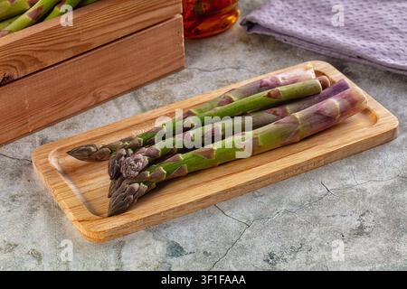 Asperges vertes fraîches avec des pointes violettes disposées sur une planche en bois clair. Caisse en bois rustique à surface en béton en arrière-plan. Un éclairage naturel met en valeur Banque D'Images
