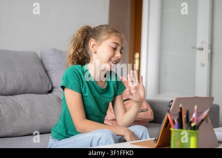 Jeune fille souriante d'école primaire faisant un signe depuis sa tablette pendant un cours en ligne depuis chez elle. Banque D'Images