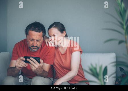 Un homme et une femme d'âge moyen sont assis côte à côte sur un canapé, lisant attentivement un message sur un smartphone. Leurs expressions traduisent de l'inquiétude, du soutien et une inquiétude partagée Banque D'Images