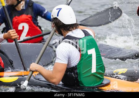 Joueur de polo en canoë masculin en chemise blanche et gilet vert pagayant dans l'eau, avec des coéquipiers en maillots rouges visibles, illustrant la dynamique sportive compétitive en Banque D'Images