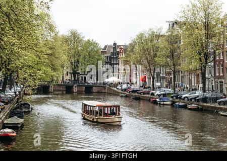 Amsterdam, pays-Bas - 18 avril 2026 : croisière touristique en bateau le long d'un canal bordé d'arbres avec des maisons hollandaises historiques dans le centre d'Amsterdam Banque D'Images
