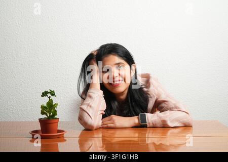 Jeune femme sourit en reposant sa tête sur la table. Sa main touche ses cheveux foncés. Une petite plante en pot se trouve à côté d'elle. La table en bois reflète sa pose. Simple Banque D'Images