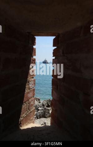Plusieurs navires, dont un patrouilleur et un grand yacht, naviguent à l'horizon, vus à travers une arche de pierre érodée du Fort Carré à Antibes Banque D'Images