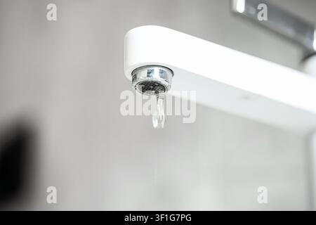 Un cliché macro détaillé montrant une seule goutte d'eau claire se formant et pendante d'un robinet blanc qui fuit, avec une accumulation visible de calcaire minéral. Banque D'Images