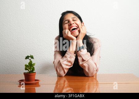Femme riant joyeusement avec les mains près du visage. Une petite plante en pot est posée à côté d'elle sur la table. Un fond simple met en valeur son expression joyeuse. Lumière chaude Banque D'Images