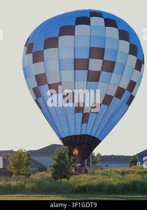 Grand ballon à air chaud à carreaux bleus et bruns atterrissant et se dégonflant dans un champ herbeux près d'un quartier de banlieue au coucher du soleil Banque D'Images