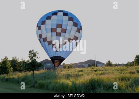 Grand ballon à air chaud à carreaux bleus et bruns atterrissant et se dégonflant dans un champ herbeux près d'un quartier de banlieue au coucher du soleil Banque D'Images