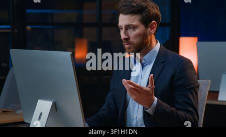 Homme d'affaires caucasien sérieux et concentré passant un appel vidéo informatique dans un bureau la nuit, homme d'affaires d'âge moyen concentré, entrepreneur, employeur Banque D'Images