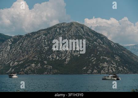 Catamaran moderne ancré sur les eaux calmes de la baie de Kotor au Monténégro. Des montagnes rocheuses et des nuages dramatiques créent un fond pittoresque de la mer Adriatique. Banque D'Images