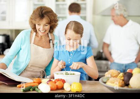 Grand-mère lisant et cuisinant avec un enfant dans la cuisine, partageant des moments ou apprenant des compétences culinaires dans l'appartement. Femme âgée heureuse et préparation du repas avec une fille Banque D'Images