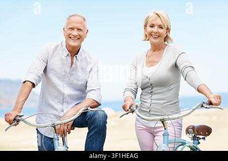 Plage, vélo et vacances avec un couple de seniors, portrait et sourire avec un moyen de transport pour voyager. Homme et femme heureux et vélo en plein air pour le cyclisme Banque D'Images