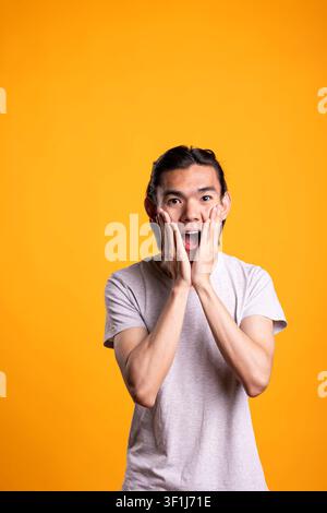 Individu masculin surpris tient ses mains aux joues avec la bouche ouverte, réagissant à des nouvelles excitantes. Jeune homme séduisant semble ravi et excité, montrant une expression joyeuse d'incrédulité et de joie. Banque D'Images