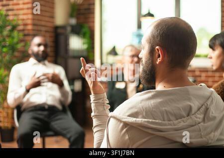 Homme barbu faisant différents signes avec les doigts pendant une séance de thérapie de groupe, jeune homme blanc assis en cercle et faisant des gestes des mains dans une pièce aux murs de briques tandis que des gens discutent en arrière-plan. Banque D'Images
