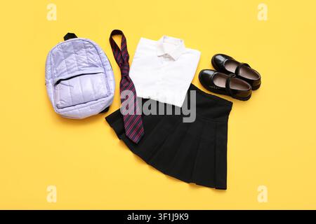 Uniforme scolaire avec chemise blanche, jupe plissée noire, cravate rayée, chaussures brillantes et sac à dos violet sur fond jaune Banque D'Images