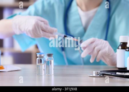 Professionnel médical en blouse et gants préparant une seringue avec un médicament provenant d’une ampoule sur une table de clinique, deux ampoules transparentes sont posées sur un bois clair Banque D'Images