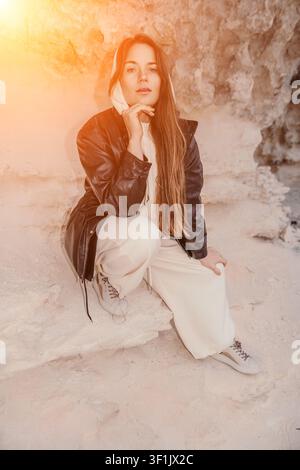 Portrait en veste en cuir d'une jeune femme en survêtement blanc posant à l'intérieur d'une grotte rocheuse baignée d'une lumière chaude de coucher de soleil Banque D'Images