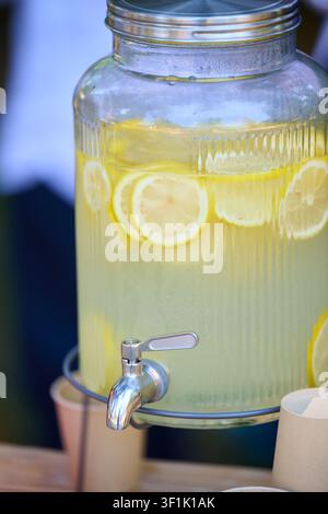 Grand distributeur en verre contenant une limonade fraîche faite maison avec des tranches de citron, prêt à être versé dans des gobelets jetables, représentant la fraîcheur et des boissons saines Banque D'Images