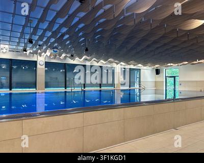 Un établissement de piscine intérieur contemporain présentant un plafond en bois ondulé frappant et une eau teintée de bleu. L'espace est propre, bien éclairé et designe Banque D'Images
