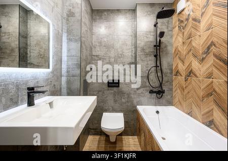 Salle de bain moderne avec murs carrelés gris, accent en bois chevron, robinets blancs et quincaillerie noire Banque D'Images