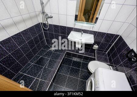 Prise de vue en hauteur d'une salle de bains carrelée comportant un lavabo, des toilettes, une douche et une machine à laver. La partie inférieure des murs et du sol est recouverte de b Banque D'Images