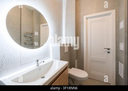 salle de bain moderne avec des carreaux gris, un miroir rond illuminé, un lavabo blanc sur une armoire en bois et une porte blanche Banque D'Images