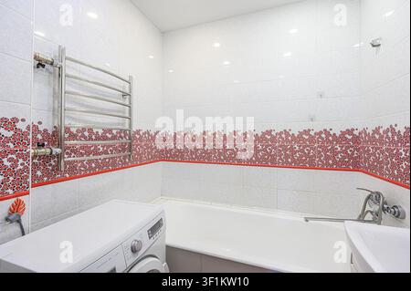 Salle de bain avec des carreaux blancs et à motifs floraux. Une machine à laver se trouve près d'une baignoire. Un porte-serviettes chauffant est fixé au mur Banque D'Images
