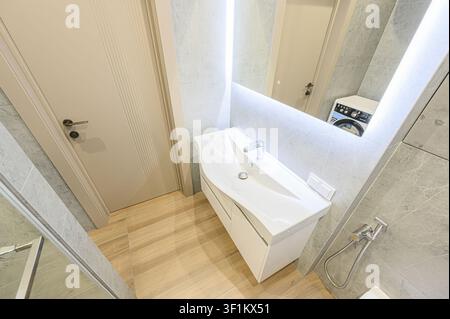 Salle de bain moderne avec un lavabo blanc, des murs en marbre gris, un miroir avec éclairage intégré et un plancher en bois Banque D'Images