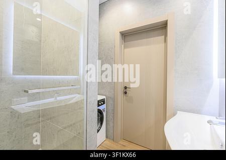 Salle de bain moderne avec carrelage en marbre, miroir éclairé, machine à laver, lavabo et une porte beige clair. Design épuré et minimaliste Banque D'Images