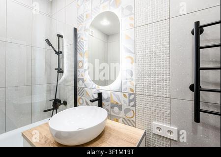 salle de bain moderne avec un lavabo ovale blanc sur un comptoir en bois, un robinet noir et des accessoires de douche, des carreaux décoratifs et un miroir éclairé Banque D'Images
