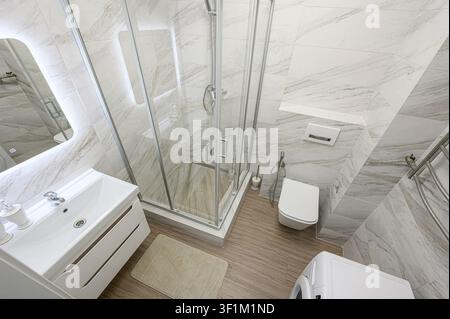 Salle de bain moderne dotée d'une douche en verre, d'un lavabo blanc, de toilettes et d'une machine à laver. L'espace est carrelé dans un motif marbré, avec un sol aspect bois Banque D'Images