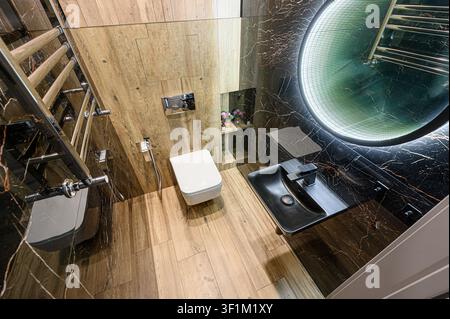 Salle de bain moderne avec carrelage aspect bois, murs en marbre noir, un miroir LED rond et des accessoires élégants Banque D'Images