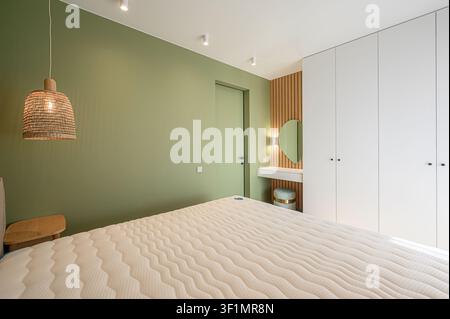 Chambre à coucher confortable avec un mur vert clair, une lampe suspendue tissée, un lit blanc et des armoires encastrées blanches. Une zone de coiffeuse moderne avec un miroir en forme de cœur Banque D'Images