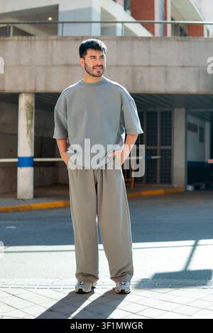 Portrait en grandeur nature d'un homme barbu élégant portant un haut gris surdimensionné et un pantalon à jambes larges, debout au soleil près d'un parking urbain, mains Banque D'Images