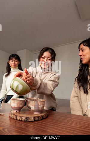 Femme japonaise souriante verse du thé chaud dans des tasses en céramique faites à la main tandis que des amis regardent, partageant un rituel du thé calme à la maison avec une vaisselle traditionnelle et une guerre Banque D'Images
