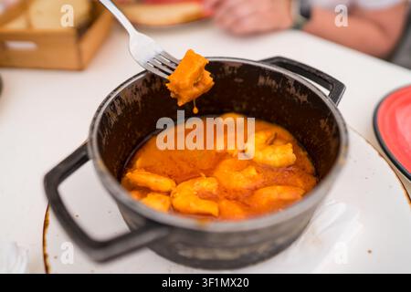 Gros plan d'une casserole noire remplie d'un ragoût de fruits de mer orange vif, comprenant des crevettes. Une fourchette soulève un morceau de ragoût. Servi avec du pain sur une table blanche dans Banque D'Images