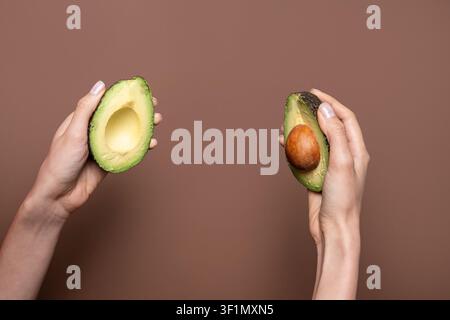 Concept alimentaire minimaliste avec deux mains tenant des moitiés d'avocat mûres, une avec le noyau, sur un fond brun chaud. Composition propre avec espace de copie pour la santé Banque D'Images
