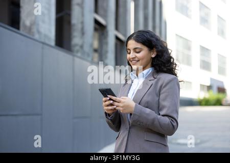Jeune femme d'affaires professionnelle souriante. Interagissant avec un smartphone tout en étant debout à l'extérieur dans un environnement urbain moderne. Transmettant des concepts de communication réussie. Mise en réseau Banque D'Images
