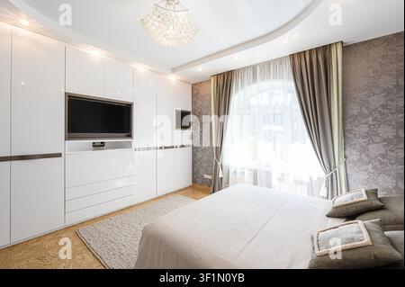 Chambre moderne dotée d'un grand lit, d'une armoire blanche brillante avec télévision intégrée et d'une fenêtre en arc avec rideaux Banque D'Images